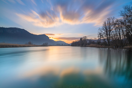 Long exposure of a colorful sunrise along the Adda cycle path. The sun rises on the horizon, coloring the clouds and the calm waters of the river with warm colours, Lecco, Lombardy, Italy