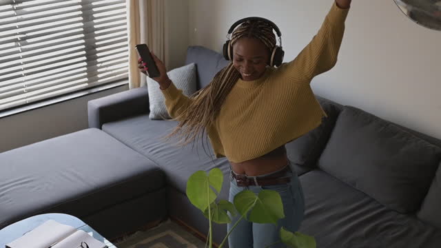 Music, black woman and dance with headphones in a living room, happy and having fun with phone playlist. Streaming, online and girl with radio, podcast or audio track in lounge, dancing and cheerful