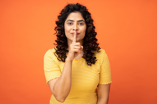 Portrait of pretty young woman showing silence sign, holding finger on lip on orange background