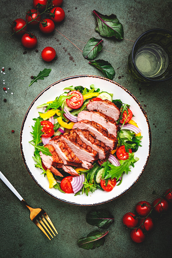Duck salad with grilled breast and vegetables: red tomato, cucumber, paprika, onoin  and chard, frisse, mizuna and arugula leaves on green table background, top view