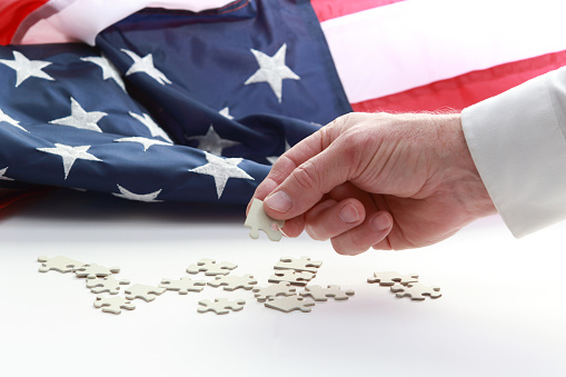 A man's hand holding white puzzle pieces, with part of the sleeve and with American flag in background.