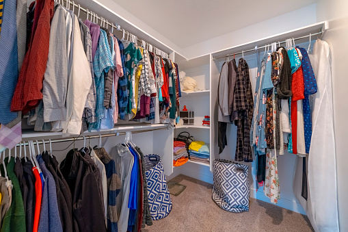 Windowless walk-in closet with corner shelves in between hanging metal rods with clothes. Walk-in closet interior with brown carpet flooring and two linen laundry basket below the shelves.