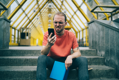 Surprised man in glasses looking in disgust at smartphone screen and holding folder in his hands while sitting on steps