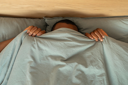 Top View Of Black Man Hiding Covering Face With Blanket Lying In Bed In Modern Bedroom At Home. Anxiety And Fear, Sleeping Routine Concept. High Angle Shot