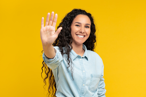 Smiling young lady outstretching hand to the camera offering giving high five or showing palm and smiling on yellow orange studio background wall, greeting someone. Hello, Nice to Meet You.