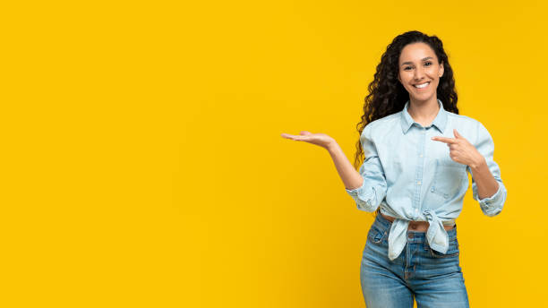 istock Smiling young woman pointing at blank space, posing and smiling over yellow studio background, panorama 1475624125