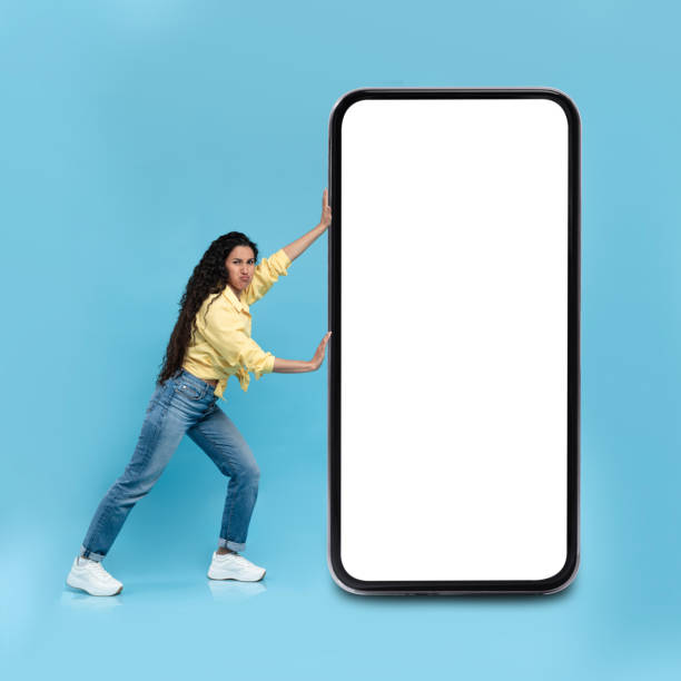 istock Emotional young woman pushing big heavy smartphone with empty screen on blue studio background, mockup 1475624221