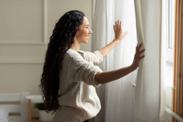 istock Smiling woman opening window curtains at home 1475624236