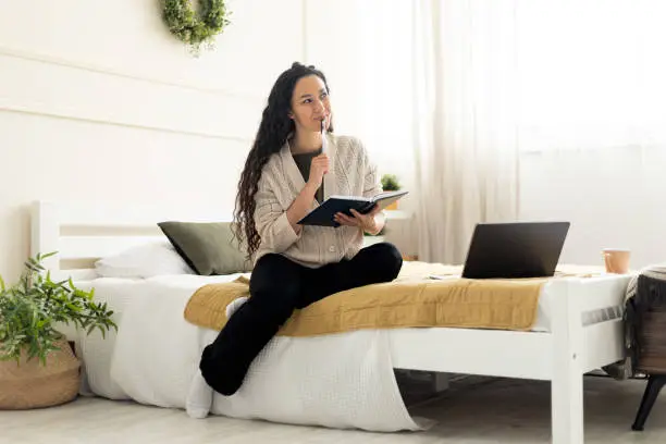 Portrait of smiling young woman holding pen writing in notebook sitting on bed with laptop computer, dreamy female looking aside and thinking, planning future, full body length, modern interior
