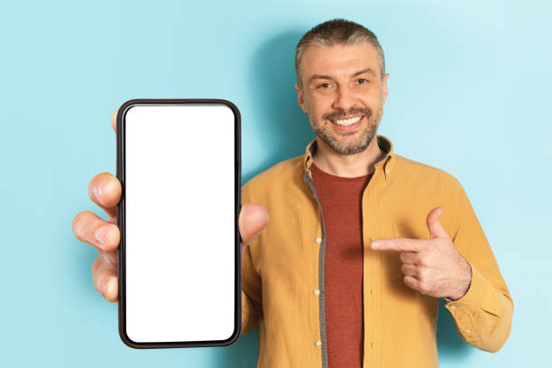 istock Mobile advertisement. Excited middle aged man pointing at cellphone with empty screen on blue background, mockup 1475624369