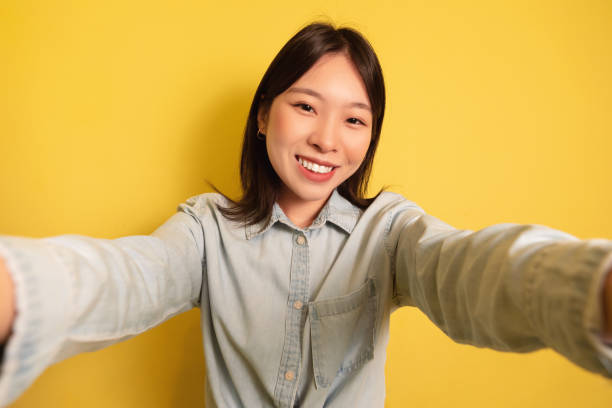 Capturing moments. Happy young Asian lady taking mobile selfie over yellow studio background
