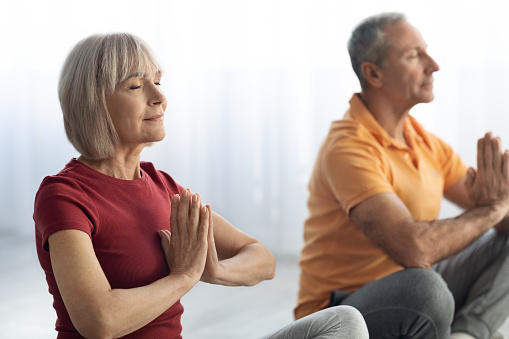 Closeup portrait of senior man and woman in sportswear practicing meditation together indoors, sitting next to each other in lotus position with closed eyes, relaxation at home, stress relief concept