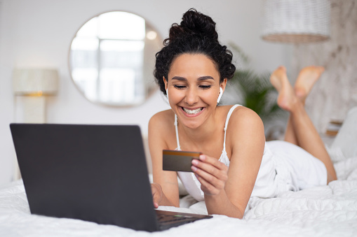 Satisfied young caucasian brunette female with curly hair in pajamas paying money with credit card and laptop on bed in bedroom interior. Online shopping and shopaholic at home due covid-19 outbreak