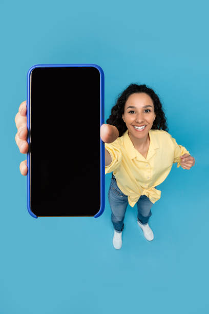 istock Pretty young lady pointing at smartphone with blank screen on blue studio background, mockup. Above view 1475624893
