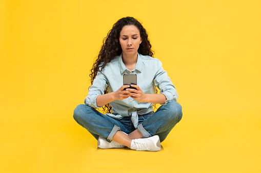 Concerned Arabic Woman Using Cellphone Texting And Frowning Reading Bad Message Sitting Over Yellow Studio Background. Mobile Communication Problems Concept