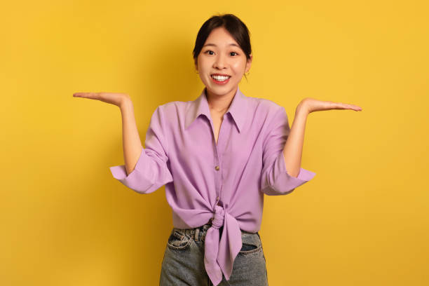 istock Happy Asian woman comparing options, making scales with hands, holding invisible objects on yellow background, mockup 1475624913