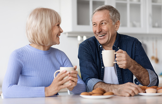 Positive beautiful elderly spouses drinking tea with pastry at home, sitting at kitchen table, holding coffee mugs, having conversation and smiling, enjoying time together, closeup