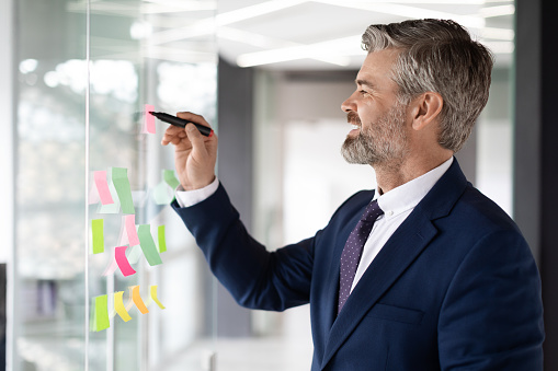 Mature Male Entrepreneur Writing On Sticky Notes On Glass Wall At Office, Smiling Middle Aged Businessman Brainstorming On New Project, Noting Business Ideas, Planning Working Strategy, Side View