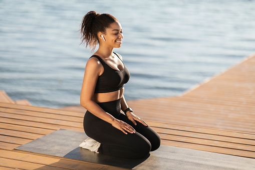 Peaceful young african american woman in sportswear meditating with closed eyes on the beach, enjoying her morning practice outdoors, sitting on fitness mat, using earbuds, side view, copy space