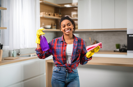 Glad millennial black woman in rubber gloves with spray, rag has fun, enjoys cleaning, use professional cleaning supplies for home perfect tidiness in kitchen interior. Household chores, housekeeping