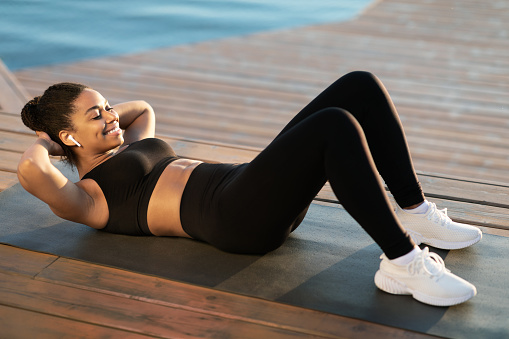 Cheerful african american young sexy lady exercising outdoors next to river, lying on yoga mat and doing crunches, working on abs and smiling, side view, copy space. Outdoor fitness concept