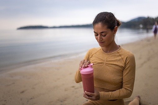 Woman on the beach by the calm sea in sunset, and holding a healthy smoothie drink.
