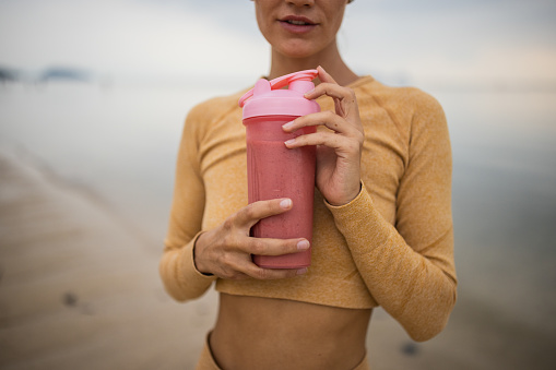 Woman on the beach by the calm sea, she is holding a healthy smoothie drink.