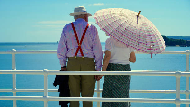 Plymouth, Devon, 06 July 2022; Images of happy days in retirement. A well dressed elderly couple enjoy the sunshine and views of the ocean. The woman uses the a colourful parasol and the gentleman a straw hat to protect themselves from the harsh rays of the sun.