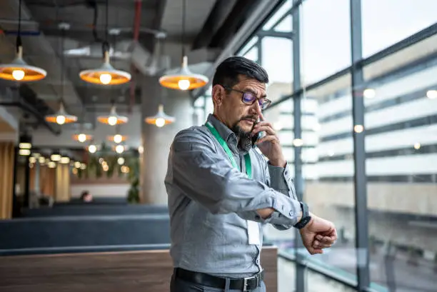 Photo of Mature man talking on the phone and checking the time on his wristwatch at office