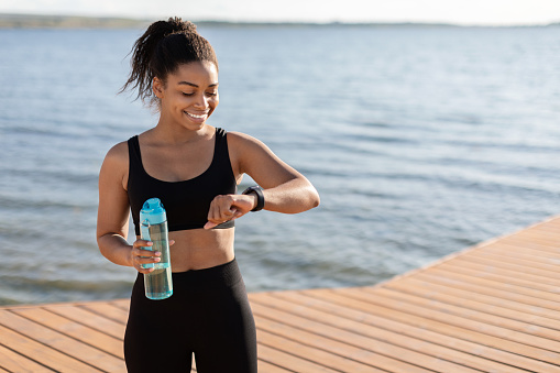 Athletic millennial black woman drinking water, checking fitness bracelet and smiling, happy african american sporty lady jogging around city lake, tracking distance and blood oxygen, copy space