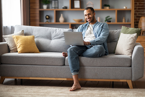 Freelance Concept. Happy Young African American Man Using Laptop At Home, Smiling Black Millennial Guy Sitting On Couch With Computer, Relaxing In Living Room With Modern Gadget, Copy Space