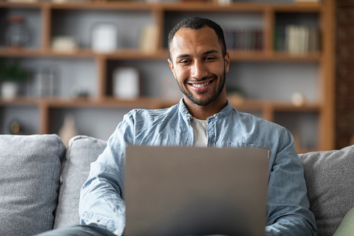 Online Communication. Handsome Young Black Man Sitting On Couch And Using Laptop, Smiling African American Guy Relaxing In Living Room At Home With Computer, Messaging In Social Networks, Closeup