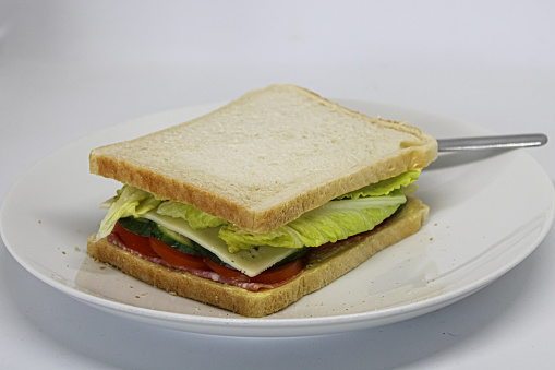 One homemade salami tomato cucumber cheese and lettuce white bread sandwich on a white plate on white background