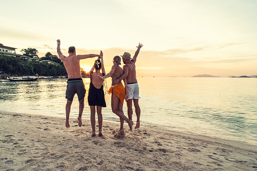 Group of happy friends enjoying beautiful sunset at the tropical beach, jumping and having fun together. Travelers. Tourism. Back view. Thailand, Asia.