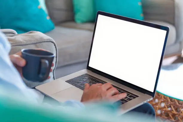 Mature man using a blank screen laptop at home. He is sitting on the sofa with a coffee.