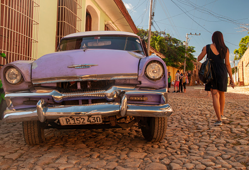 Trinidad, Cuba - December 21, 2017: A purple vintage car on the cobblestone streets of Cuba with a woman walking by
