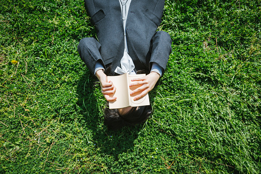 Asian teenage girl wearing school uniform lying down on grass and reading a book.