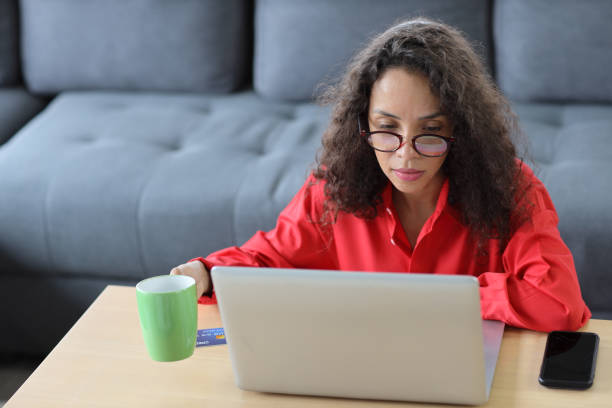 istock Smart and active latin woman sitting and using internet on computer with smart mobile phone in living room. Lifestyle with technology 1475692967