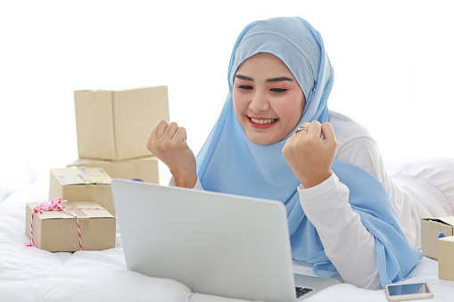 Beautiful and young asian woman in muslim sleepwear with attractive look, lies on bed with computer, mobile phone and online package box delivery. Smart girl with hijab receive good news and surprise.