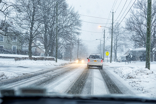 Car driver's point of view of winter blizzard snow storm vehicle traffic moving slowly along a slippery, slushy suburban village street. A yellow school crossing sign with walking school children symbols is brightly illuminated up ahead.