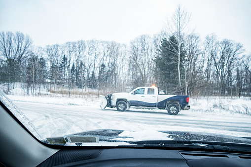 Winter snowplow pick-up truck passing across a rural road intersection in February. Car driver point of view through a frozen car windshield.