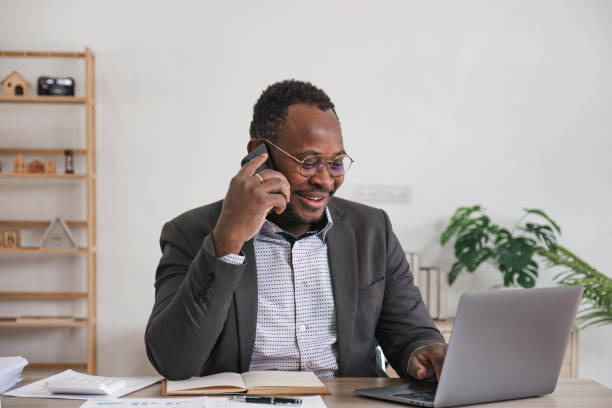 istock Happy black businessman talking on cellphone having phone conversation sitting at workplace working on laptop computer in office. 1475728845