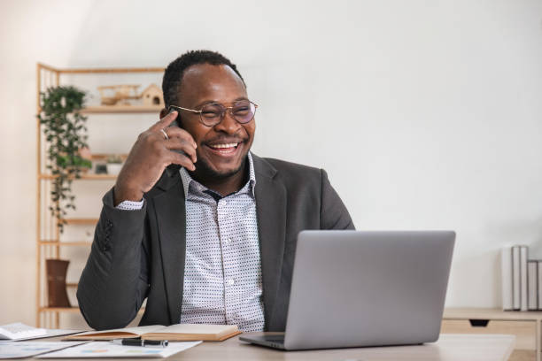 istock Happy black businessman talking on cellphone having phone conversation sitting at workplace working on laptop computer in office. 1475728855