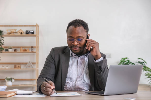istock Happy black businessman talking on cellphone having phone conversation sitting at workplace working on laptop computer in office. 1475728866