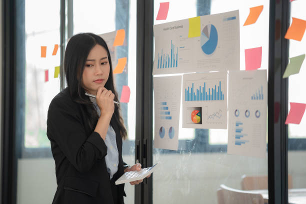 istock Young Asian businesswoman working on a sticky note on board, planning and analyzing business finance and marketing at the office. 1475730297
