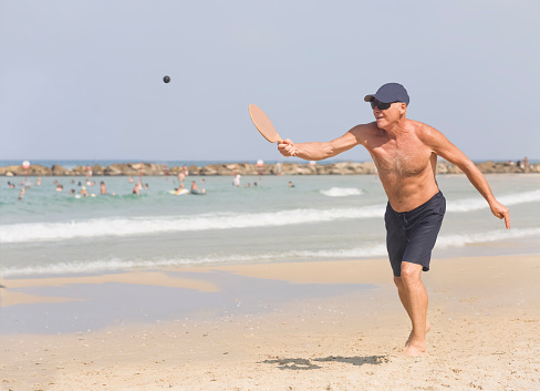 A handsome mature man hits a ball while playing matkot on the beach. Active life in retirement. Full-height. Weekend in Israel