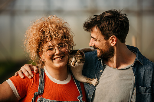 Close up portrait of a cheerful embraced diverse couple of workers standing in their greenhouse, playing with a cat.