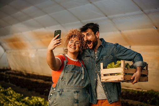 Caucasian friendly woman and man farmer smiling, taking a selfie, and holding organic fresh green vegetables produce wooden box together in greenhouse garden nursery farm, business farmer and healthy food concept.