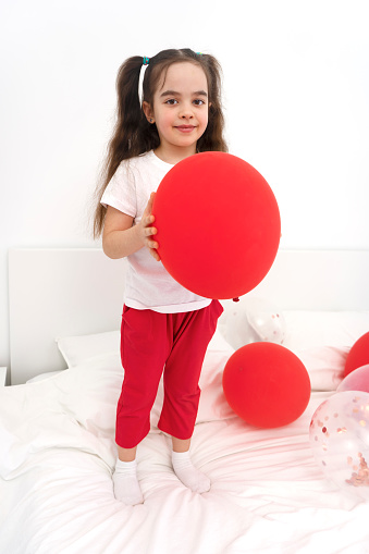 Positive emotions preschooler kid 6-7 years old standing on the bed in the bedroom and holding red balloons. Birthday celebration party. Happy childhood.