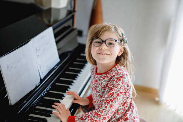 Cтоковое фото Маленькая счастливая девочка играет на пианино в гостиной. Милый дошкольник с очками развлекается с обучением игре на музыкальном инструм�